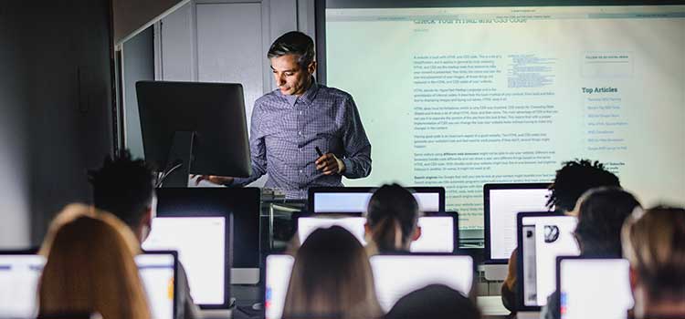 professor at front of class teaching room of students on laptops